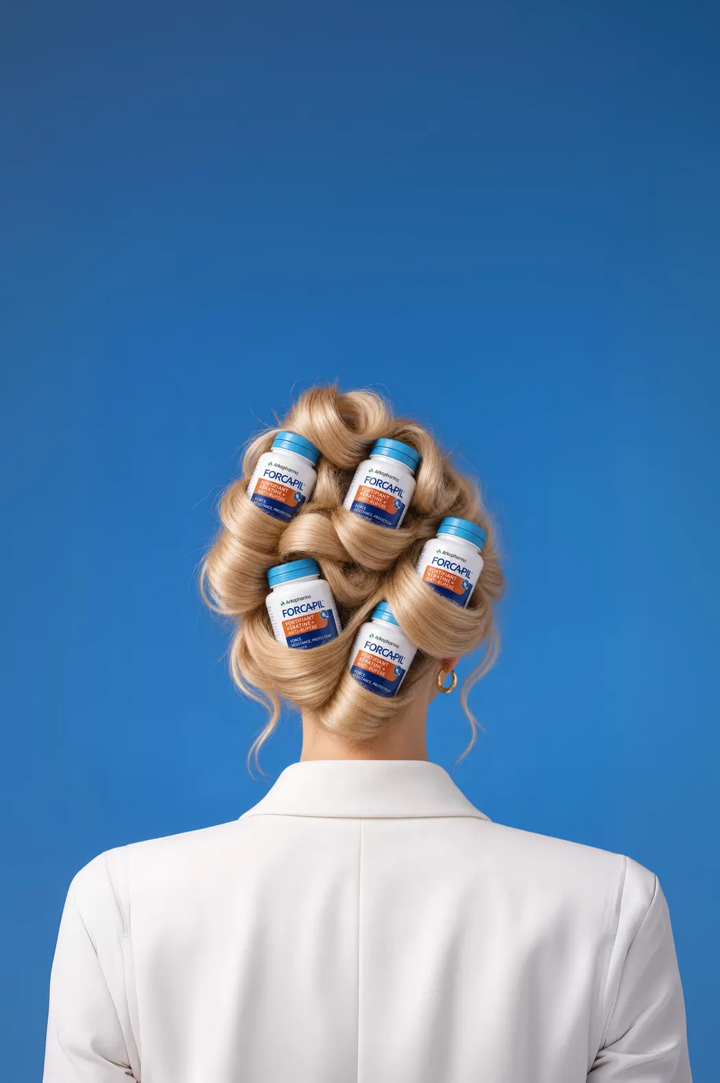 Femeie blondă, văzută din spate, cu părul prins în coc în care sunt așezate flacoane FORCAPIL® Fortifiant Keratine+